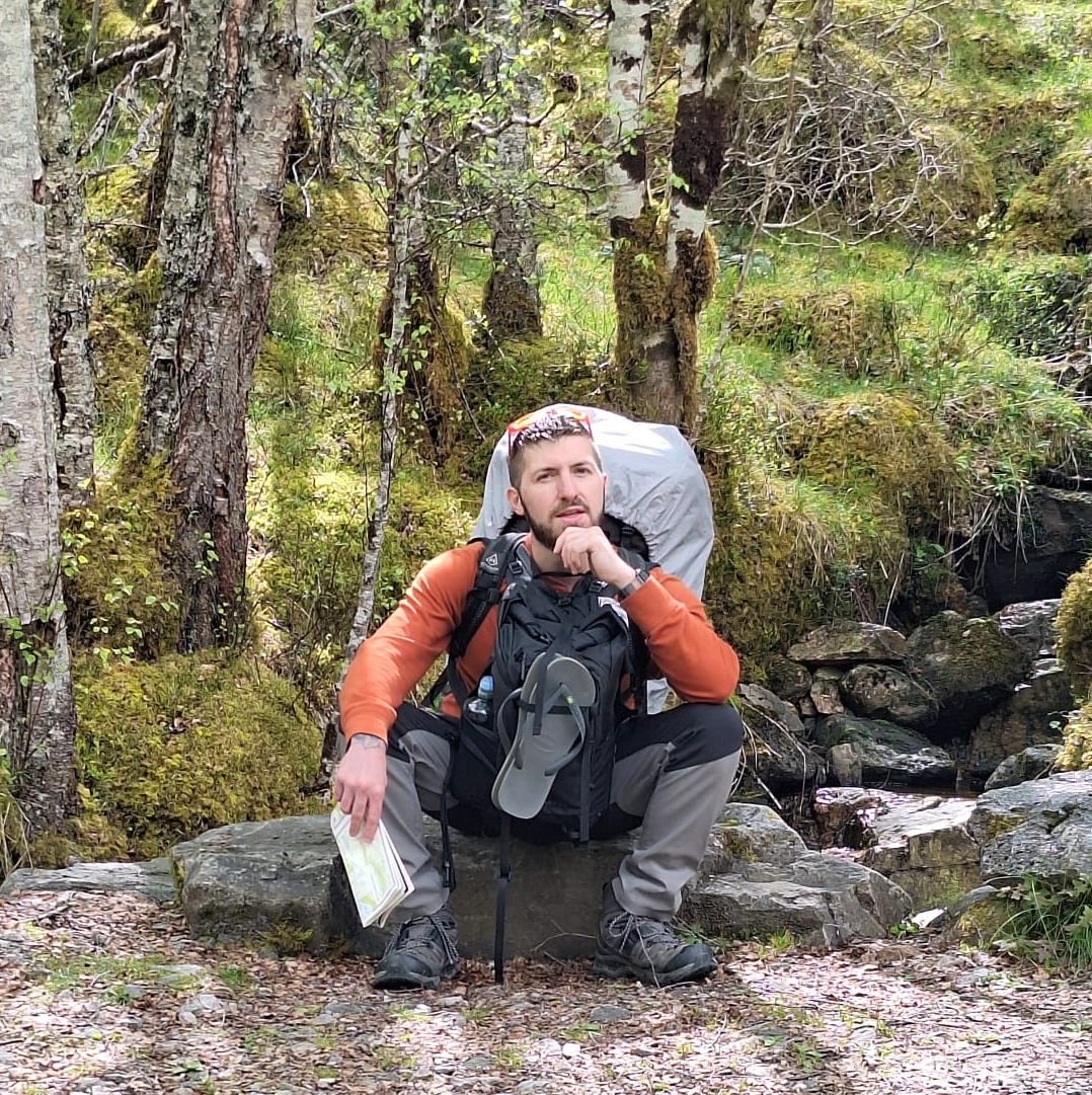 Brandon Shewmake seated on rocks beside a small stream in a wooded mountain setting, wearing hiking gear and carrying a pack.